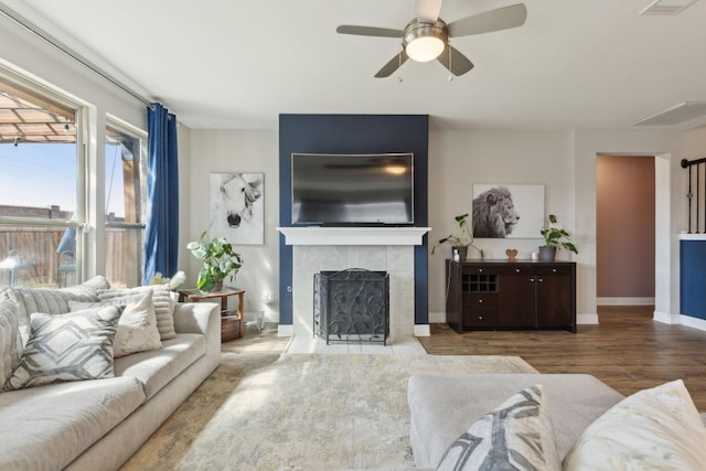 living area with ceiling fan, a tiled fireplace, wood finished floors, and baseboards