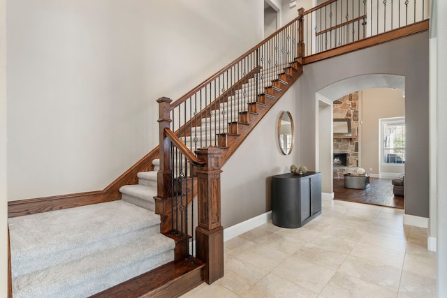 staircase featuring arched walkways, baseboards, a stone fireplace, and a high ceiling