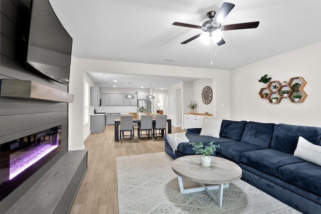 living area with a glass covered fireplace, visible vents, ceiling fan, and light wood-style flooring