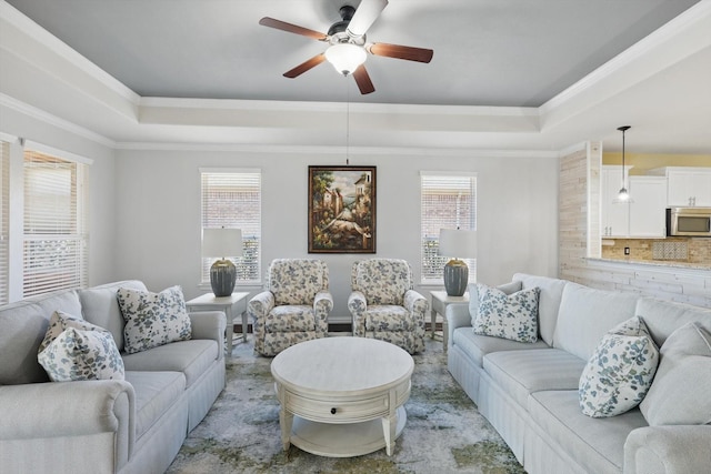 living room with a healthy amount of sunlight, ornamental molding, a raised ceiling, and a ceiling fan