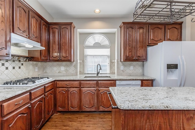 kitchen featuring light stone counters, under cabinet range hood, white appliances, a sink, and dark wood finished floors