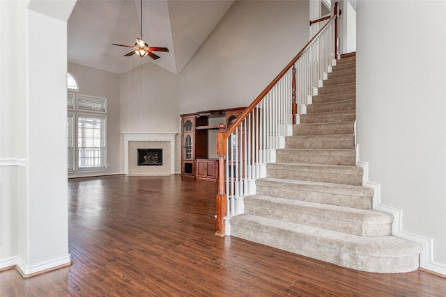 staircase featuring high vaulted ceiling, a premium fireplace, wood finished floors, a ceiling fan, and baseboards