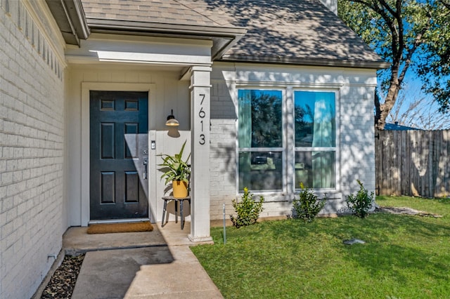 view of exterior entry with a shingled roof, fence, and a yard