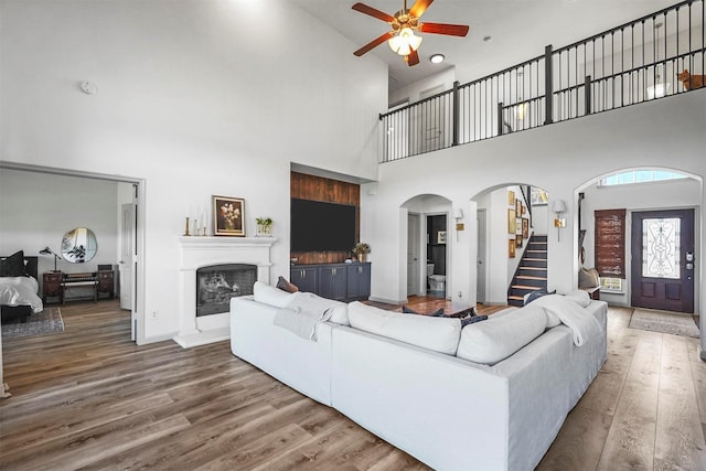 living room featuring arched walkways, a fireplace with raised hearth, stairway, ceiling fan, and wood finished floors