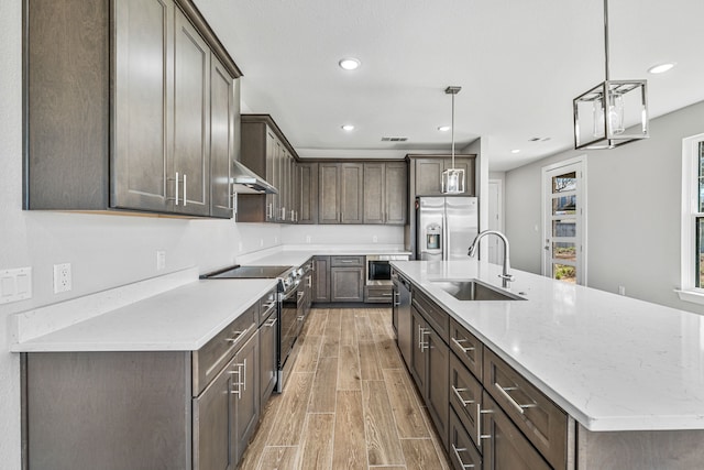 kitchen featuring an island with sink, appliances with stainless steel finishes, wood finish floors, wall chimney range hood, and a sink