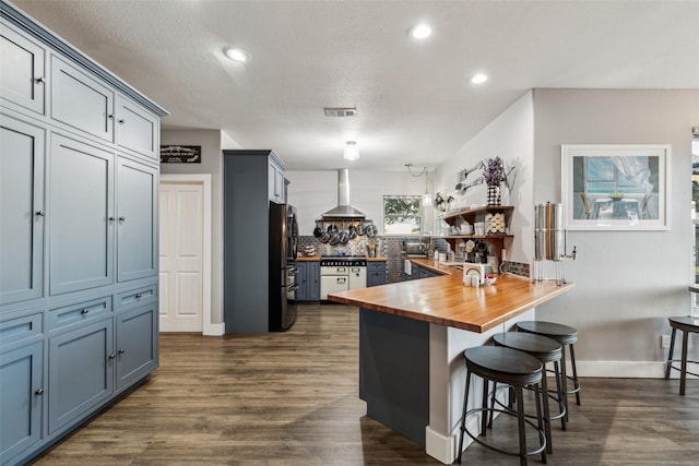 kitchen with a breakfast bar area, a peninsula, wooden counters, freestanding refrigerator, and wall chimney exhaust hood