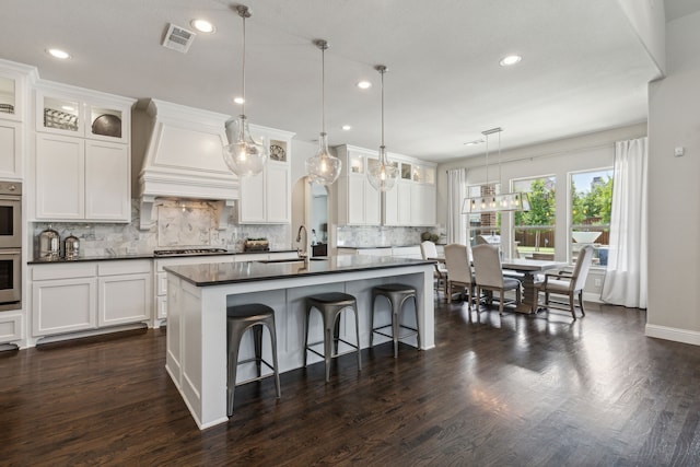 kitchen with visible vents, dark countertops, an island with sink, premium range hood, and white cabinetry