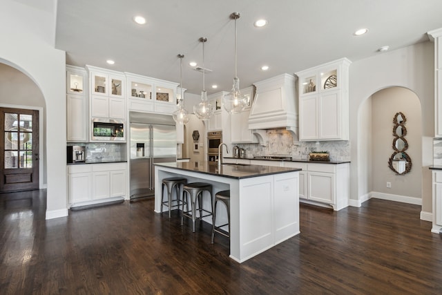 kitchen featuring dark wood-style floors, arched walkways, dark countertops, built in appliances, and premium range hood