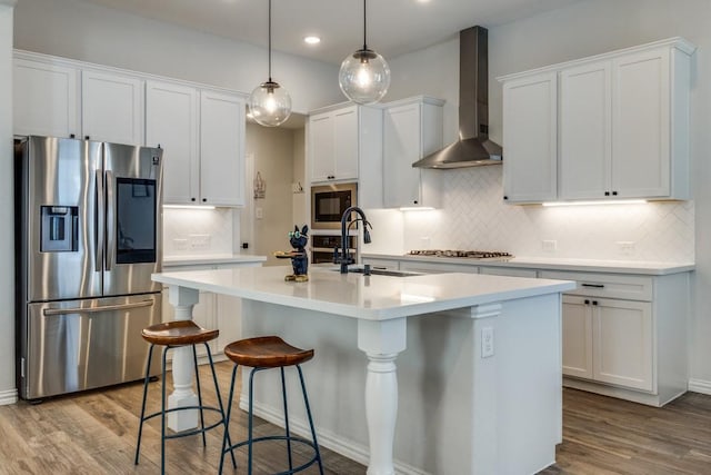 kitchen with a kitchen island with sink, stainless steel appliances, wood finished floors, a sink, and wall chimney range hood