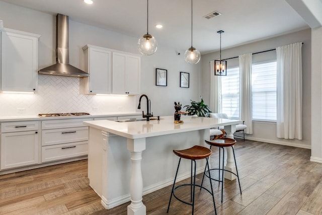 kitchen with light countertops, backsplash, a sink, wall chimney range hood, and an island with sink