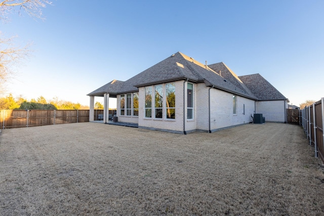 rear view of house with central air condition unit, a fenced backyard, brick siding, and roof with shingles