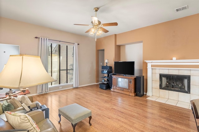 living room with visible vents, a tiled fireplace, a ceiling fan, wood finished floors, and baseboards