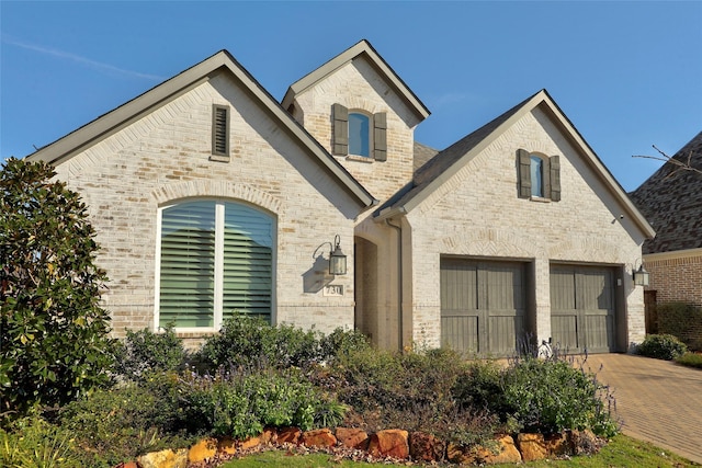 french country home with a garage, brick siding, and decorative driveway