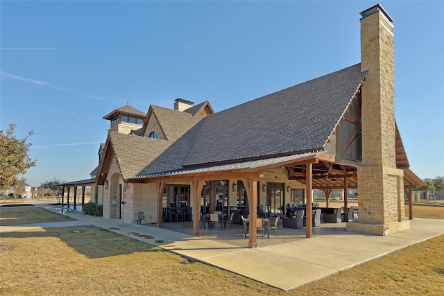 back of house with a shingled roof, a patio, a chimney, and a lawn