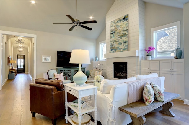 living area with a fireplace, high vaulted ceiling, light wood-type flooring, baseboards, and ceiling fan with notable chandelier