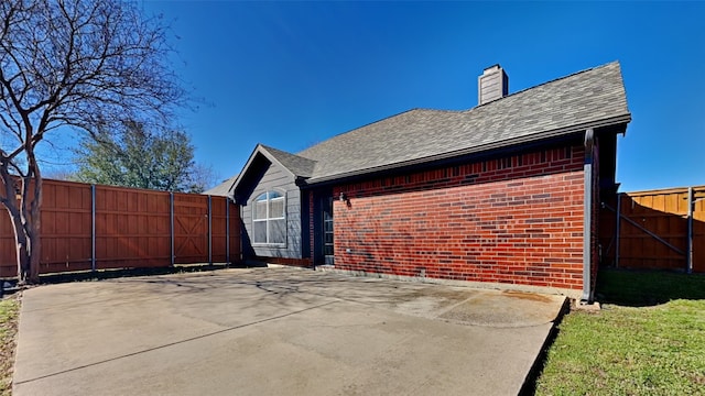 view of side of home with roof with shingles, brick siding, a chimney, and fence