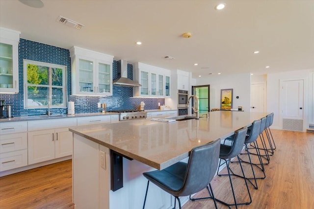 kitchen featuring visible vents, a large island with sink, wall chimney range hood, and light wood finished floors