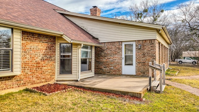 exterior space with brick siding, a yard, a chimney, roof with shingles, and a patio area