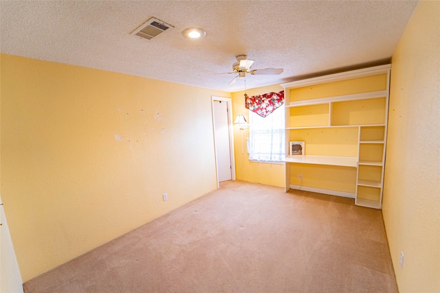 unfurnished room featuring a textured ceiling, light colored carpet, visible vents, a ceiling fan, and built in desk