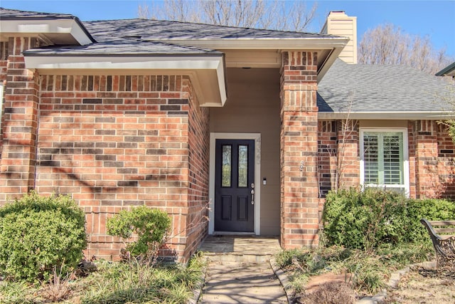 view of exterior entry with roof with shingles, a chimney, and brick siding