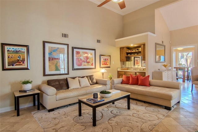 living room with a high ceiling, a ceiling fan, visible vents, baseboards, and ornamental molding