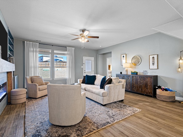 living area featuring a glass covered fireplace, ceiling fan, a textured ceiling, wood finished floors, and baseboards