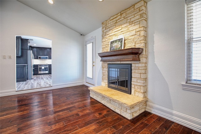 unfurnished living room featuring lofted ceiling, arched walkways, a stone fireplace, baseboards, and hardwood / wood-style floors