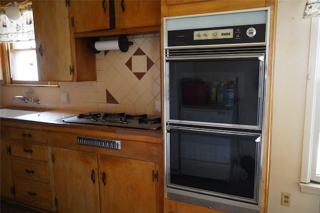 kitchen featuring tile countertops, a sink, multiple ovens, stainless steel gas cooktop, and backsplash