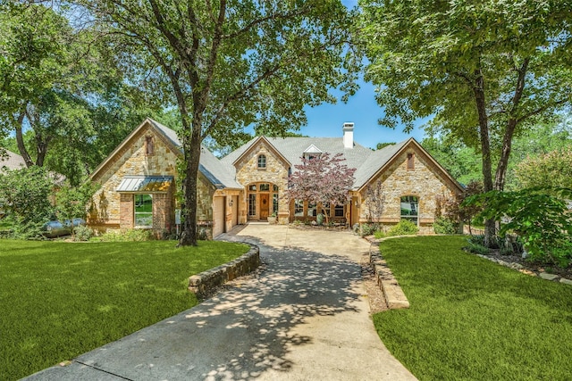 french country inspired facade featuring concrete driveway, stone siding, a chimney, an attached garage, and a front yard