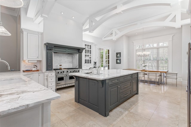 kitchen with an island with sink, light stone counters, a sink, white cabinets, and range with two ovens