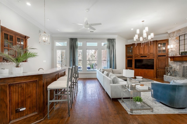 living room with ceiling fan with notable chandelier, crown molding, a stone fireplace, and dark wood-style floors