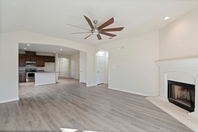 unfurnished living room featuring arched walkways, a fireplace with raised hearth, a ceiling fan, and light wood-style floors