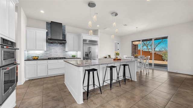 kitchen with stainless steel appliances, white cabinets, backsplash, wall chimney exhaust hood, and a center island with sink