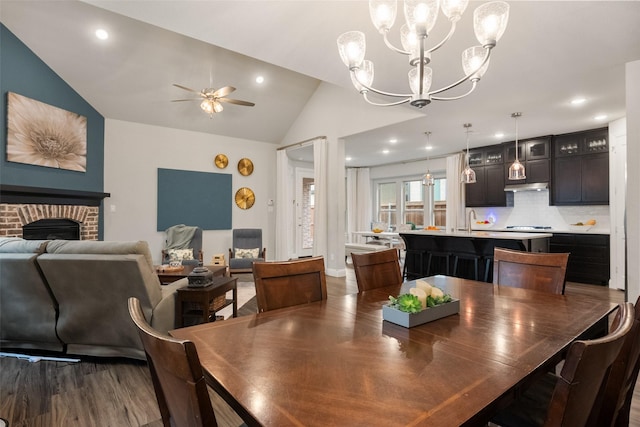 dining space with wood finished floors, ceiling fan with notable chandelier, vaulted ceiling, a brick fireplace, and recessed lighting