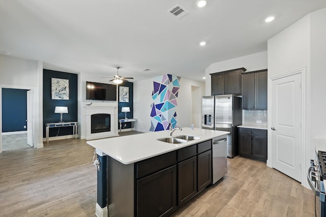 kitchen with a center island with sink, stainless steel appliances, light countertops, a glass covered fireplace, and a sink