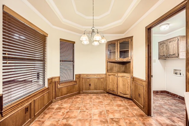 unfurnished dining area with a wainscoted wall, a raised ceiling, a chandelier, and crown molding