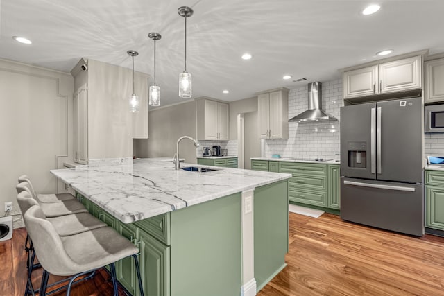 kitchen featuring a breakfast bar, a sink, appliances with stainless steel finishes, green cabinets, and wall chimney range hood