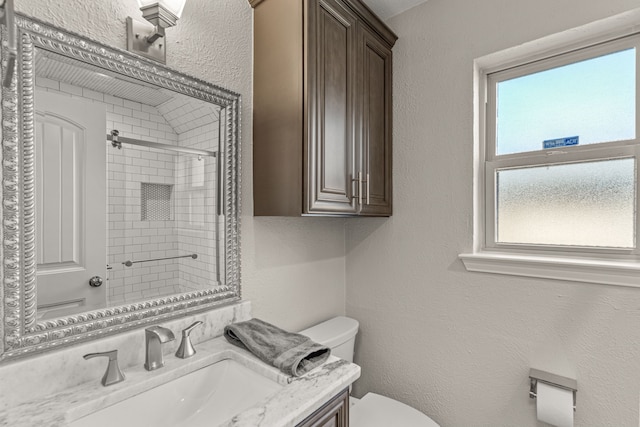 bathroom featuring tiled shower, toilet, vanity, and a textured wall