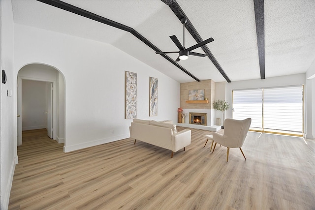 living area featuring ceiling fan, a textured ceiling, vaulted ceiling with beams, light wood-style flooring, and a large fireplace