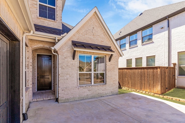 view of exterior entry featuring roof with shingles, fence, a patio, and brick siding