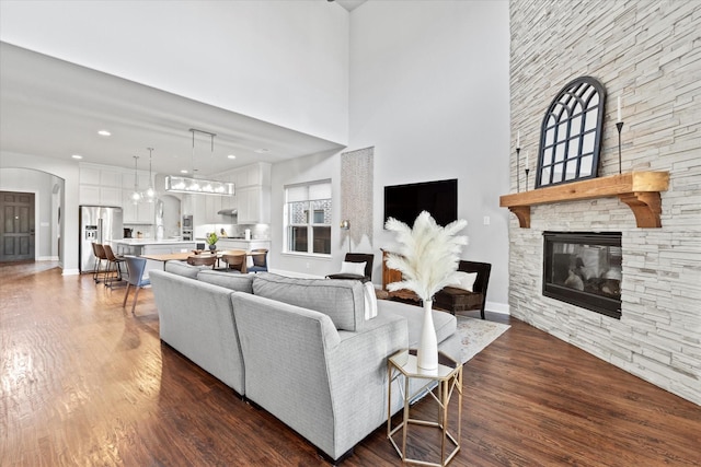 living area featuring arched walkways, a stone fireplace, a towering ceiling, baseboards, and dark wood-style floors