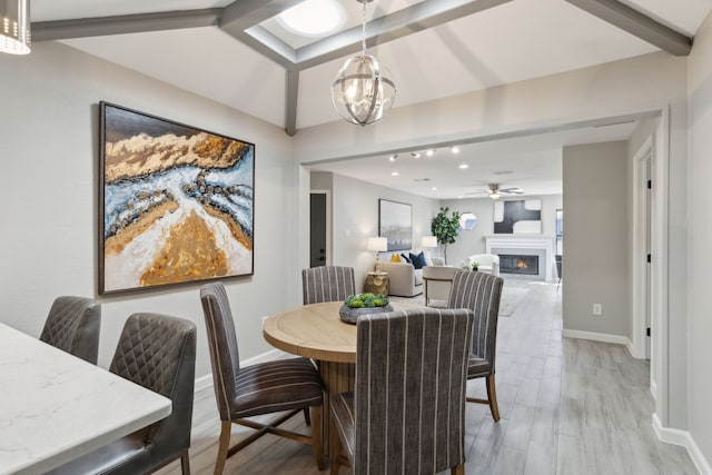 dining room with ceiling fan with notable chandelier, baseboards, a lit fireplace, beam ceiling, and light wood finished floors