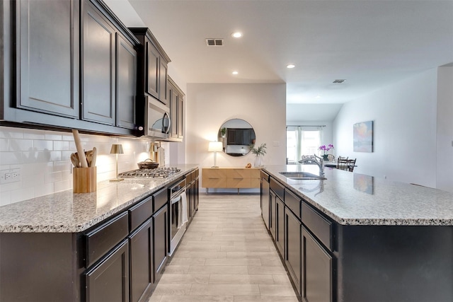 kitchen with light stone counters, a kitchen island with sink, a sink, appliances with stainless steel finishes, and backsplash