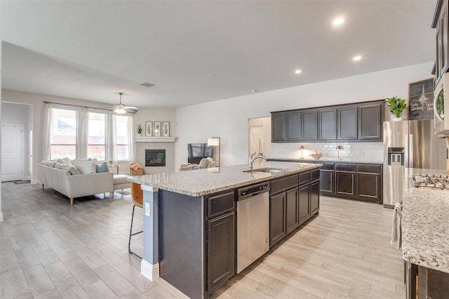 kitchen featuring a center island with sink, stainless steel appliances, backsplash, a sink, and dark brown cabinets