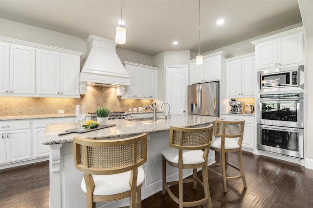 kitchen with a center island with sink, dark wood-style floors, stainless steel appliances, premium range hood, and a sink