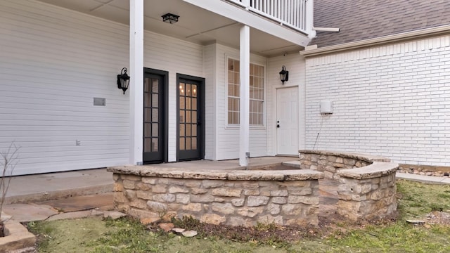 property entrance featuring covered porch, roof with shingles, brick siding, and a balcony