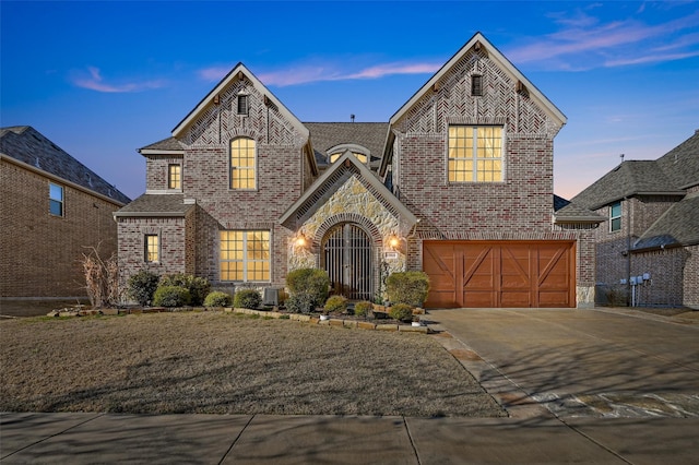 french country inspired facade with a garage, a shingled roof, concrete driveway, stone siding, and brick siding