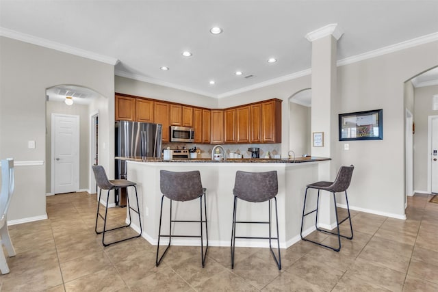 kitchen featuring arched walkways, decorative backsplash, appliances with stainless steel finishes, brown cabinetry, and a kitchen bar