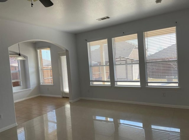 tiled empty room with arched walkways, visible vents, a ceiling fan, a textured ceiling, and baseboards