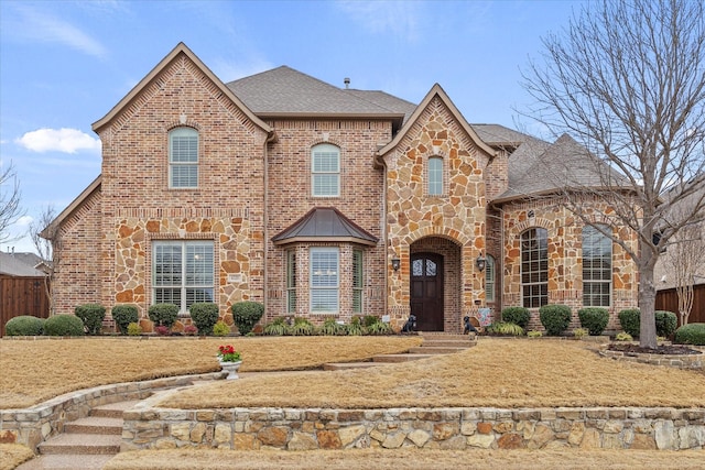 french country inspired facade with stone siding, brick siding, a shingled roof, and fence